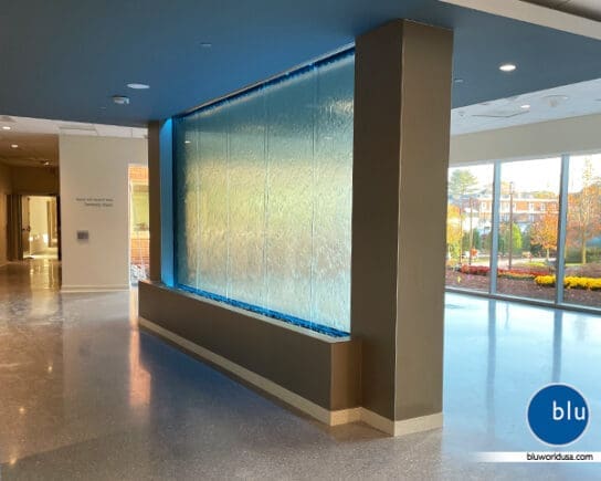 This encapsulated horizontal water feature has five panels of clear glass with color changing light at the top and bottom and located in between the walkway and waiting area of a hospital.